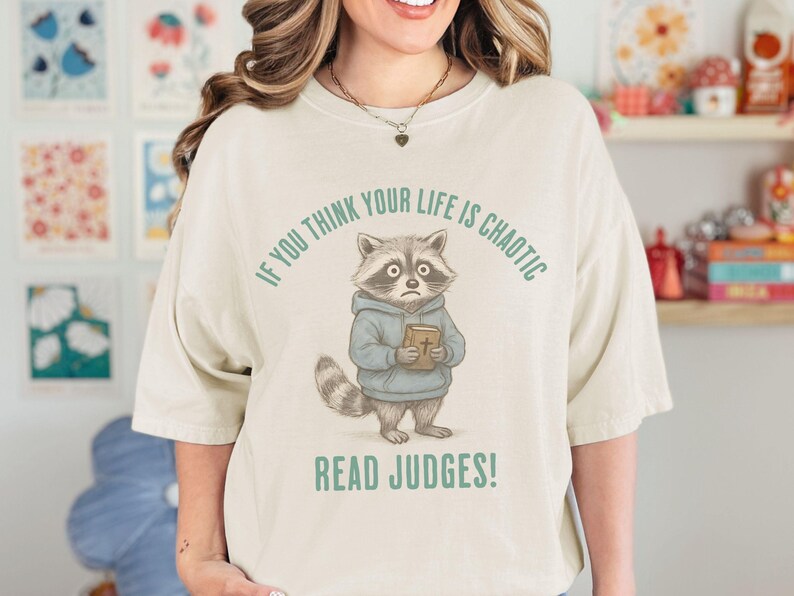 May include: Cream-colored t-shirt featuring a cartoon raccoon wearing a blue hoodie and holding a book. The text "IF YOU THINK YOUR LIFE IS CHAOTIC" arches above the raccoon, with "READ JUDGES!" below.
