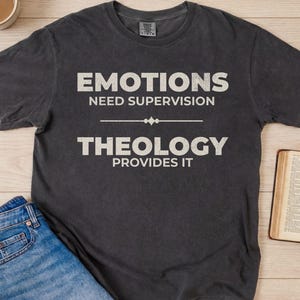 May include: Dark gray t-shirt with the text "EMOTIONS NEED SUPERVISION THEOLOGY PROVIDES IT" in white. The shirt is paired with blue jeans and an open book. A cup of coffee sits in the top left corner.