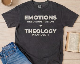 Op de afbeelding: Donkergrijs T-shirt met de tekst "EMOTIONS NEED SUPERVISION THEOLOGY PROVIDES IT" in het wit. Het shirt wordt gecombineerd met een blauwe jeans en een open boek. Een kopje koffie staat in de linkerbovenhoek.