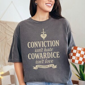 Op de afbeelding: Een donkergrijs t-shirt met de tekst "CONVICTION isn't hate, COWARDICE isn't love" en een zwaard- en hartafbeelding. Het shirt bevat ook het bijbelvers Efezi&euml;rs 4:15. Een geblokte oranje en witte mok wordt in de hand gehouden.
