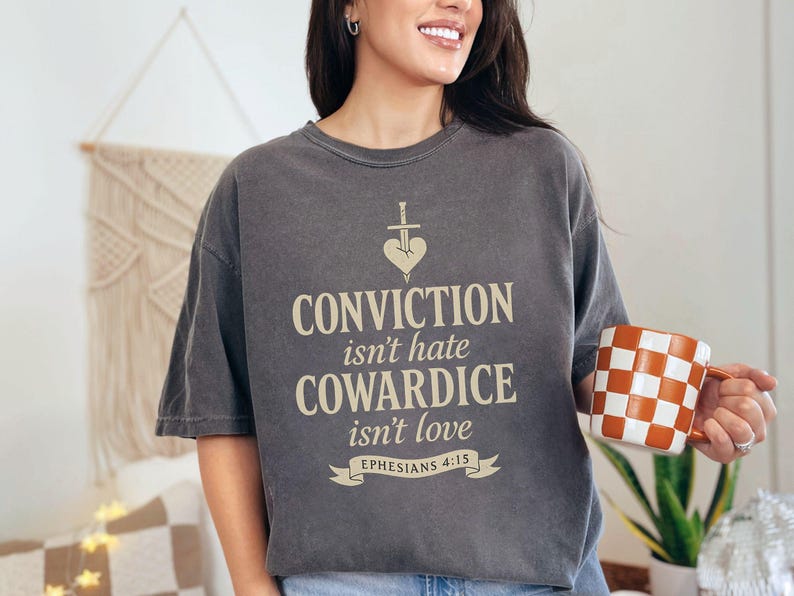 Op de afbeelding: Een donkergrijs t-shirt met de tekst "CONVICTION isn't hate, COWARDICE isn't love" en een zwaard- en hartafbeelding. Het shirt bevat ook het bijbelvers Efezi&euml;rs 4:15. Een geblokte oranje en witte mok wordt in de hand gehouden.
