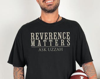 Op de afbeelding: Zwart T-shirt met de tekst "REVERENCE MATTERS ASK UZZAH" in een vetgedrukte schreefletter. Het shirt is donker van kleur en lijkt van een zacht materiaal te zijn. Een voetbal wordt in de handen van de persoon gehouden.
