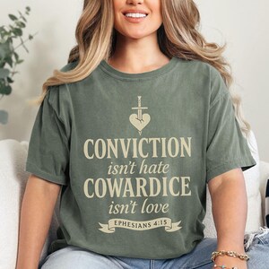 Op de afbeelding: Olijfgroen T-shirt met de tekst "CONVICTION isn't hate COWARDICE isn't love EPHESIANS 4:15". Het ontwerp bevat een hart doorboord met een zwaard. Het shirt heeft een relaxte pasvorm.