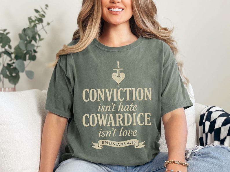 Op de afbeelding: Olijfgroen T-shirt met de tekst "CONVICTION isn't hate COWARDICE isn't love EPHESIANS 4:15". Het ontwerp bevat een hart doorboord met een zwaard. Het shirt heeft een relaxte pasvorm.
