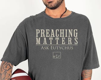 May include: Dark gray t-shirt with the text "PREACHING MATTERS" above "Ask Eutychus". The shirt has a relaxed fit and a vintage look. A football is visible in the lower portion of the image.