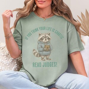 May include: Sage green t-shirt featuring a cartoon raccoon wearing a blue hoodie and holding a book. The text above the raccoon reads "IF YOU THINK YOUR LIFE IS CHAOTIC" and below it reads "READ JUDGES!"