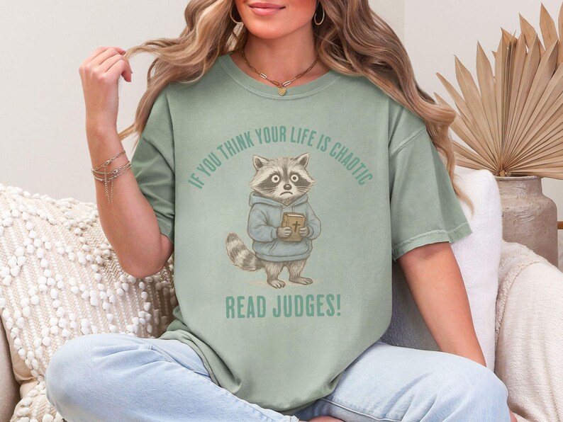 May include: Sage green t-shirt featuring a cartoon raccoon wearing a blue hoodie and holding a book. The text above the raccoon reads "IF YOU THINK YOUR LIFE IS CHAOTIC" and below it reads "READ JUDGES!"
