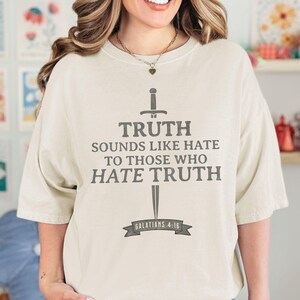 May include: Cream-colored t-shirt with the text "TRUTH SOUNDS LIKE HATE TO THOSE WHO HATE TRUTH" in gray, along with a sword graphic. The text "GALATIANS 4:16" is below the text. The shirt is a casual style.