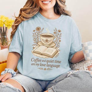 May include: Light blue t-shirt with a graphic of a coffee cup, open book, and floral elements. The text "Coffee and quiet time are my love language" is printed below the graphic. The person is holding a blue and white checkered mug.