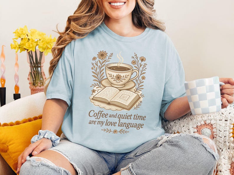 May include: Light blue t-shirt with a graphic of a coffee cup, open book, and floral elements. The text "Coffee and quiet time are my love language" is printed below the graphic. The person is holding a blue and white checkered mug.