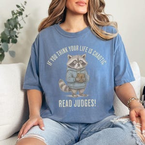 May include: A light blue t-shirt with a raccoon graphic holding a book. The text on the shirt reads, "If you think your life is chaotic, read Judges!" The raccoon is wearing a blue hoodie.
