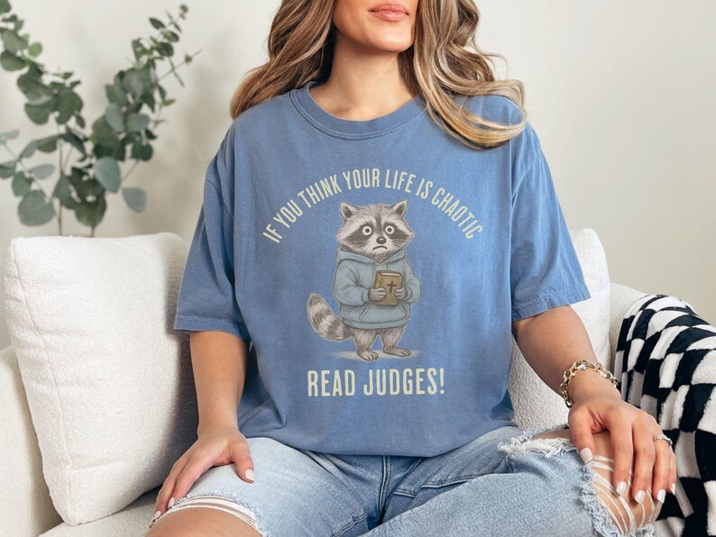 May include: A light blue t-shirt with a raccoon graphic holding a book. The text on the shirt reads, "If you think your life is chaotic, read Judges!" The raccoon is wearing a blue hoodie.