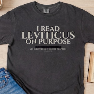 May include: Dark grey t-shirt with the text "I READ LEVITICUS ON PURPOSE" in white. Below the main text is "YES EVEN THE SKIN DISEASE CHAPTERS" and "2 Timothy 1:6". The shirt is paired with blue jeans and an open book.