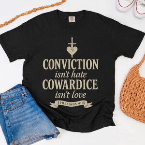 May include: Black t-shirt with the text "CONVICTION isn't hate COWARDICE isn't love" and a heart-shaped design with a sword. The shirt is paired with blue denim shorts, a woven orange bag, and white sneakers.