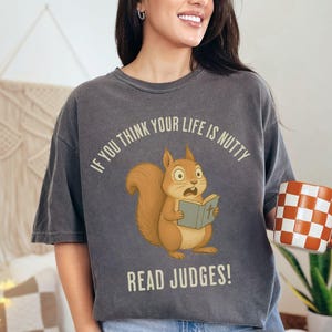 May include: Dark gray t-shirt featuring a cartoon squirrel reading a book, with the text "IF YOU THINK YOUR LIFE IS NUTTY READ JUDGES!". A checkered orange and white mug is held in the hand.