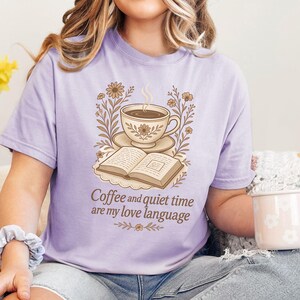 May include: A light purple t-shirt with a graphic of a coffee cup, open book, and floral accents. The text "Coffee and quiet time are my love language" is printed below the graphic. The person is wearing blue jeans and holding a white mug.