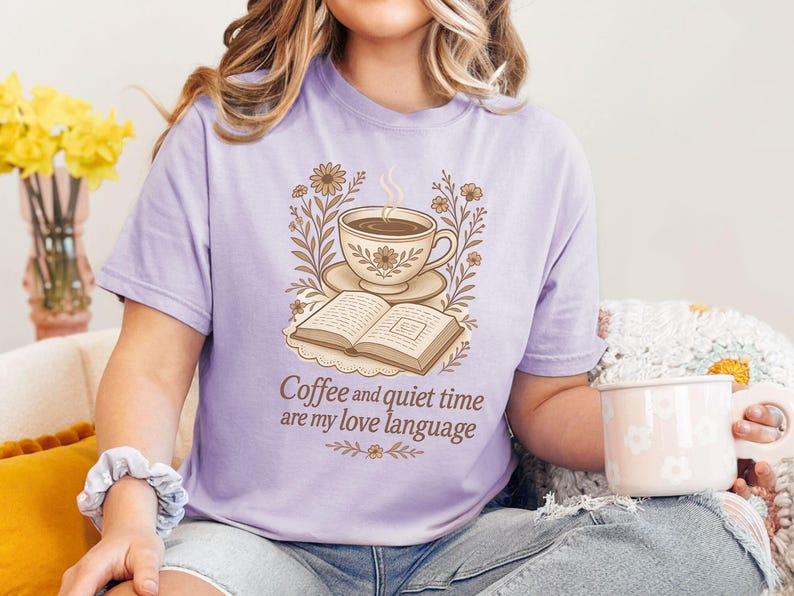May include: A light purple t-shirt with a graphic of a coffee cup, open book, and floral accents. The text "Coffee and quiet time are my love language" is printed below the graphic. The person is wearing blue jeans and holding a white mug.