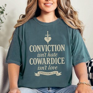 Op de afbeelding: Een tealkleurig t-shirt met de tekst "CONVICTION isn't hate, COWARDICE isn't love" in een lichtbeige lettertype. Het shirt heeft een hart- en zwaardafbeelding boven de tekst en de verwijzing "Efezi&euml;rs 4:15" eronder.