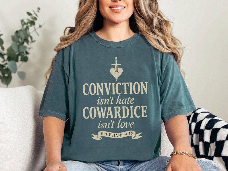 Op de afbeelding: Een tealkleurig t-shirt met de tekst "CONVICTION isn't hate, COWARDICE isn't love" in een lichtbeige lettertype. Het shirt heeft een hart- en zwaardafbeelding boven de tekst en de verwijzing "Efezi&euml;rs 4:15" eronder.