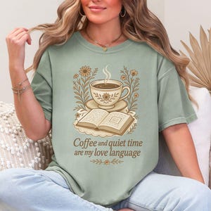 May include: Sage green t-shirt featuring a coffee cup, open book, and floral design. The text "Coffee and quiet time are my love language" is printed below the graphic. The shirt has a relaxed fit.