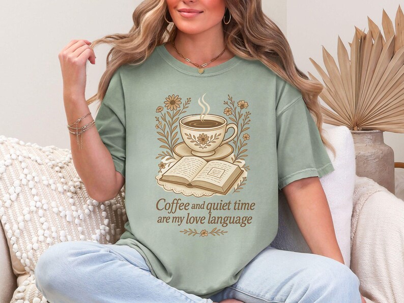 May include: Sage green t-shirt featuring a coffee cup, open book, and floral design. The text "Coffee and quiet time are my love language" is printed below the graphic. The shirt has a relaxed fit.