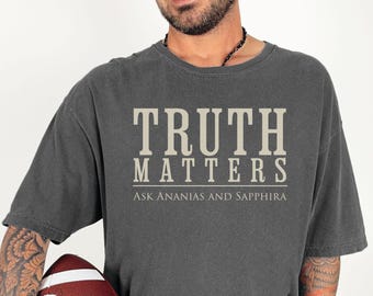 Op de afbeelding: Donkergrijs t-shirt met de tekst "TRUTH MATTERS" in een schreefletter, met daaronder "ASK ANANIAS AND SAPPHIRA". De persoon draagt het t-shirt en houdt een voetbal vast.