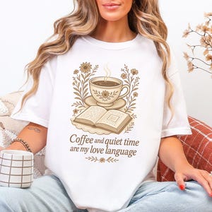 May include: White t-shirt with a sepia-toned graphic of a coffee cup, saucer, and open book, surrounded by floral elements. The text "Coffee and quiet time are my love language" is below the graphic. The person is holding a mug.
