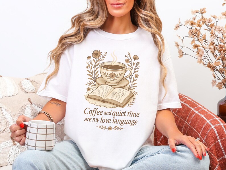 May include: White t-shirt with a sepia-toned graphic of a coffee cup, saucer, and open book, surrounded by floral elements. The text "Coffee and quiet time are my love language" is below the graphic. The person is holding a mug.