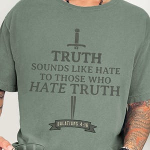 May include: Sage green t-shirt with the text "TRUTH SOUNDS LIKE HATE TO THOSE WHO HATE TRUTH" in dark gray, along with a sword graphic. The text "GALATIANS 4:16" is below the sword. The shirt is a comfortable, casual style.