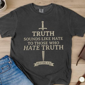 May include: Dark gray t-shirt with the text "TRUTH SOUNDS LIKE HATE TO THOSE WHO HATE TRUTH" and a sword graphic. The text "GALATIANS 4:16" is below. The shirt is laid on a wooden surface with jeans, a coffee cup, a book, and a watch.