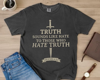 Op de afbeelding: Donkergrijs t-shirt met de tekst "TRUTH SOUNDS LIKE HATE TO THOSE WHO HATE TRUTH" en een zwaardafbeelding. De tekst "GALATIANS 4:16" staat eronder. Het shirt ligt op een houten oppervlak met een spijkerbroek, een koffiekopje, een boek en een horloge.