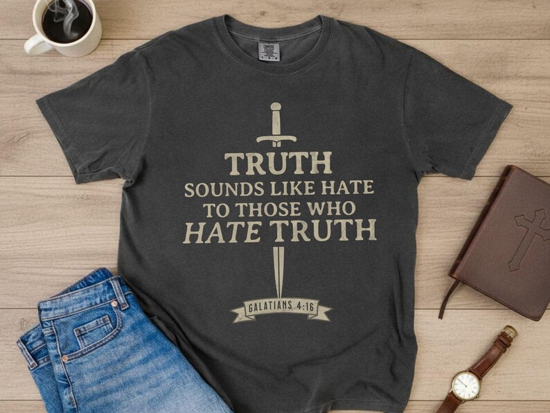 May include: Dark gray t-shirt with the text "TRUTH SOUNDS LIKE HATE TO THOSE WHO HATE TRUTH" and a sword graphic. The text "GALATIANS 4:16" is below. The shirt is laid on a wooden surface with jeans, a coffee cup, a book, and a watch.