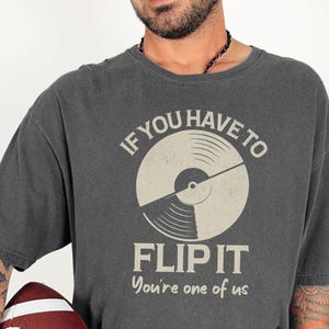 Puede incluir: Camiseta gris oscuro con un gráfico de un disco de vinilo y el texto "IF YOU HAVE TO FLIP IT You're one of us." La camiseta es de estilo vintage y está hecha de un material suave. La persona sostiene un balón de fútbol americano.