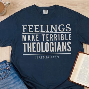 May include: Navy blue t-shirt with the text "FEELINGS MAKE TERRIBLE THEOLOGIANS" and "JEREMIAH 17:9" in white. The shirt is laid flat with a pair of blue jeans, a cup of coffee, and an open book.