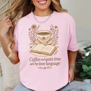 May include: Pink t-shirt with a graphic of a coffee cup, saucer, and open book, surrounded by floral elements. The text "Coffee and quiet time are my love language" is below the image.