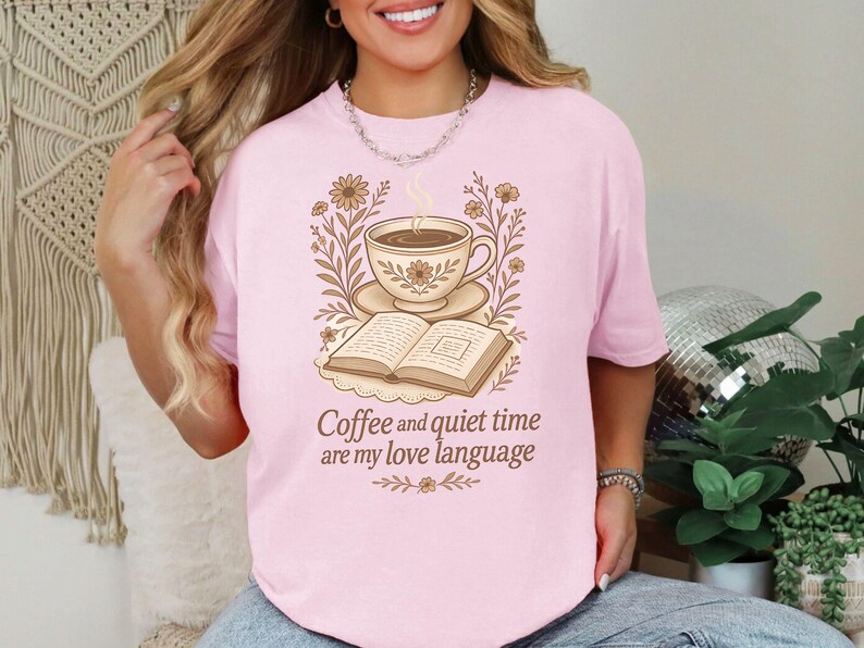 May include: Pink t-shirt with a graphic of a coffee cup, saucer, and open book, surrounded by floral elements. The text "Coffee and quiet time are my love language" is below the image.
