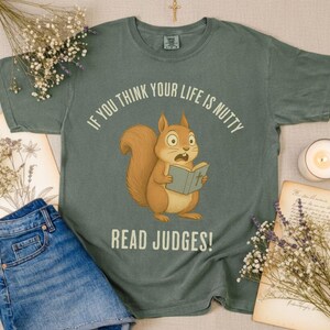 May include: Sage green t-shirt with a cartoon squirrel reading a book, and the text "IF YOU THINK YOUR LIFE IS NUTTY READ JUDGES!". The shirt is displayed with denim shorts, dried flowers, and a lit candle.