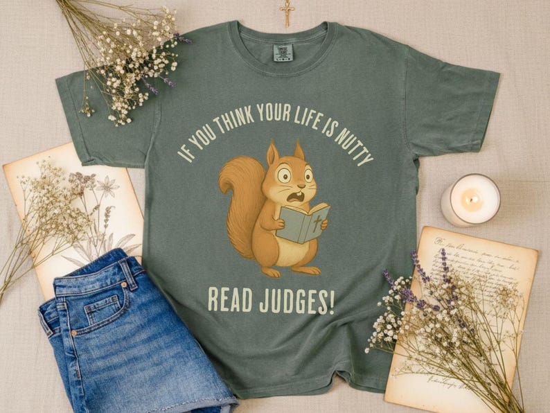 May include: Sage green t-shirt with a cartoon squirrel reading a book, and the text "IF YOU THINK YOUR LIFE IS NUTTY READ JUDGES!". The shirt is displayed with denim shorts, dried flowers, and a lit candle.