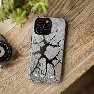 May include: A light gray phone case with a cracked design and the name "Robertson" printed at the bottom. The case has a textured surface and a black crack pattern. The phone case is on a wooden surface.