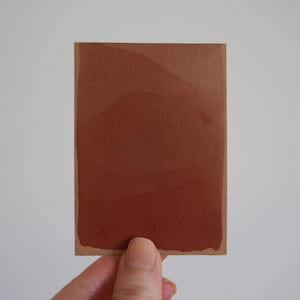 Op de afbeelding: Een rechthoekige kaart met een solide blok diepbruine terracottakleur in het midden, waarbij een dunne, onbeschilderde lichtbruine rand zichtbaar blijft langs de randen van het papier. De verfaanbrenging vertoont subtiele variaties.