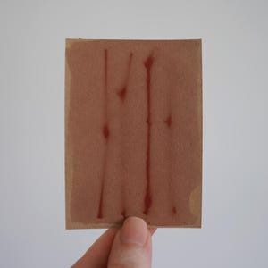 Op de afbeelding: Een rechthoekig stuk beige karton met drie verticale, dunne, roodbruin geschilderde lijnen die van de bovenrand tot de onderrand lopen, met kleine, onregelmatige, donkerbruine verfdruppels zichtbaar langs de lijnen.