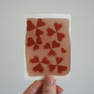 May include: A rectangular white card featuring a soft, muted brown background with a repeating pattern of dark red, three-leaf clover shapes. The clover motifs are arranged in a scattered layout across the surface of the card.