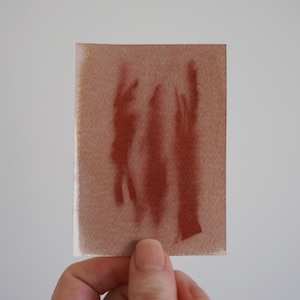 May include: A rectangular piece of textured paper featuring three vertical, abstract brushstrokes in a deep terracotta red colour. The paper has a light tan, fibrous appearance, and the paint application shows variations in pigment density.