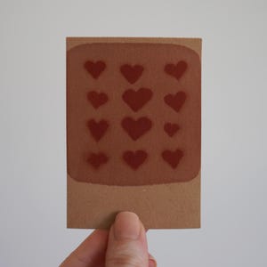 May include: A rectangular tan card featuring a centered, darker brown block with rounded corners. Inside this block are twelve dark red heart shapes arranged in a grid pattern of four rows and three columns, with the center heart being larger.