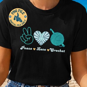 Op de afbeelding: Een zwart t-shirt met een grafisch ontwerp van een vredesteken, een hart gemaakt van haaksteken en een bol garen met een haaknaald. De tekst luidt Peace Love Crochet, gescheiden door kleine gele hartjes. Inclusief een digitaal bestand-badge.