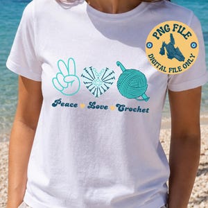 Op de afbeelding: Een wit t-shirt met een grafisch ontwerp van een turquoise vredesteken, een hart gemaakt van haaksteken en een bol wol met een haaknaald. Onder de afbeelding staat de tekst Peace, Love, Crochet met kleine gele hartjes tussen de woorden.