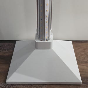 May include: A white, square-based LED grow light with a silver and white light fixture. The light fixture is cylindrical and contains multiple small LED lights. The base is a white, square shape with a pyramidal design.