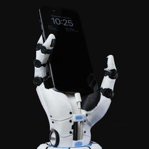 May include: A white 3D printed robotic hand holding a black smartphone with a digital clock displaying 10:25. The hand is attached to a white base with a blue ring.