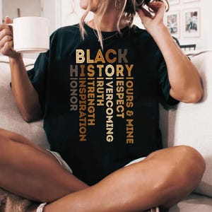 May include: Black t-shirt with the words "Black History" in gold lettering. The words "Ours & Mine", "Respect", "Overcoming", "Truth", "Strength", "Inspiration", "Honor" are stacked vertically in gold lettering.