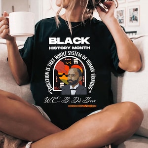 May include: Black t-shirt with a graphic design featuring a portrait of W.E.B. Du Bois and the quote "Education is that whole system of human training." The text "Black History Month" is at the top of the design.